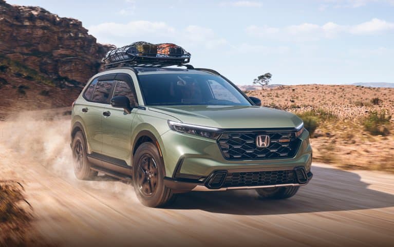 2027 Honda CR-V TrailSport Hybrid in Ash Green Metallic front driving on a dusty desert road (Compact Crossover SUV).